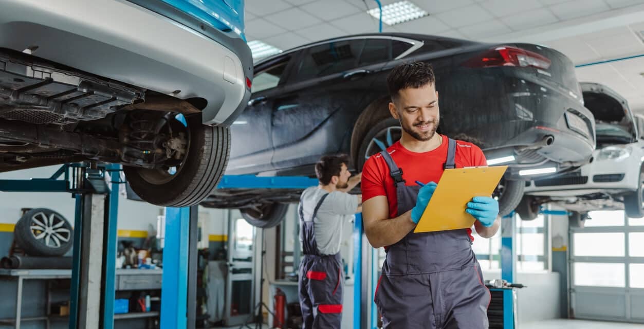 A car mechanic in a vehicle service centre completes a service checklist while another mechanic works on a car raised on jacks in the background