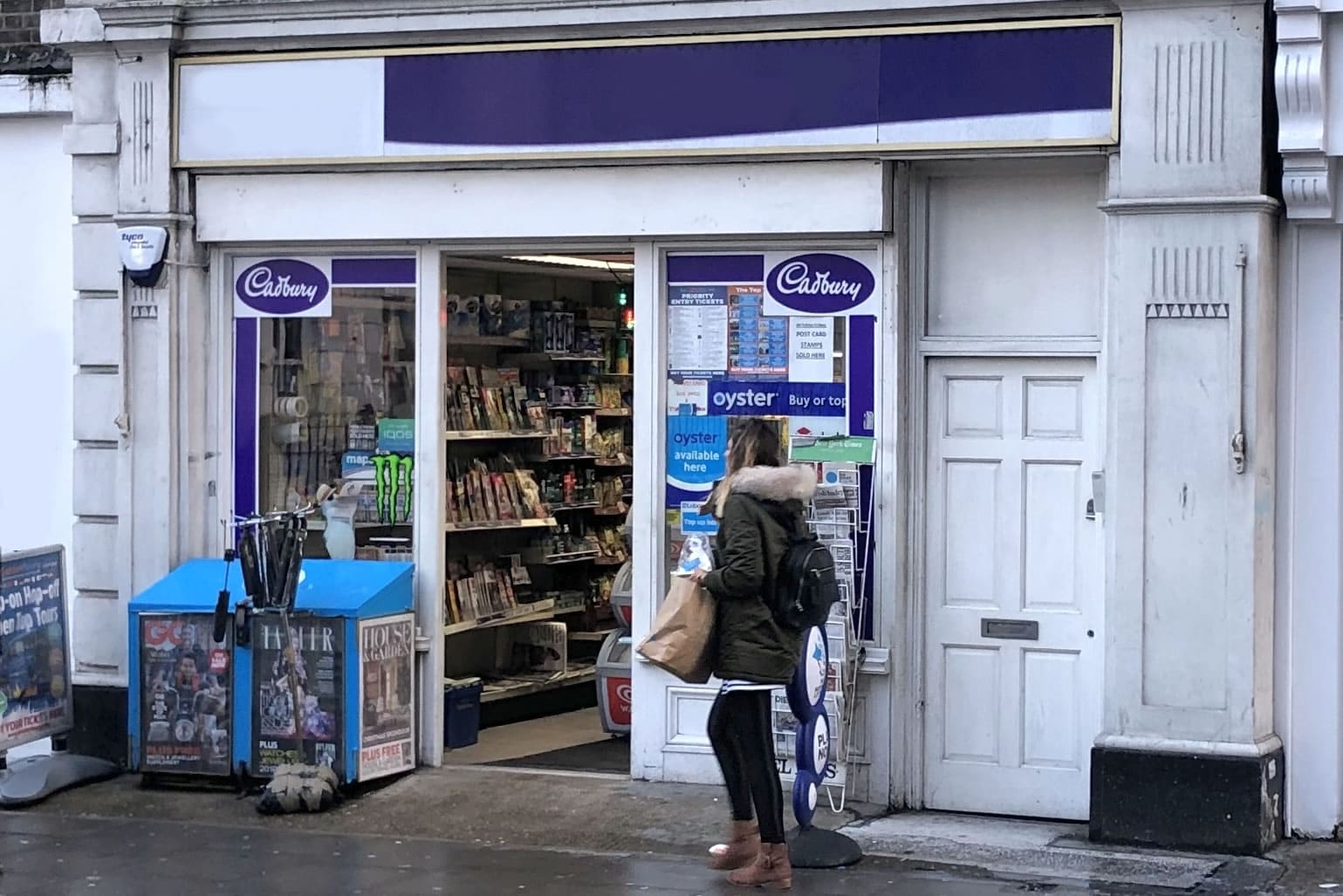 Young female consumer walking past a London corner shop selling SIMs and mobile top-ups on a rainy day
