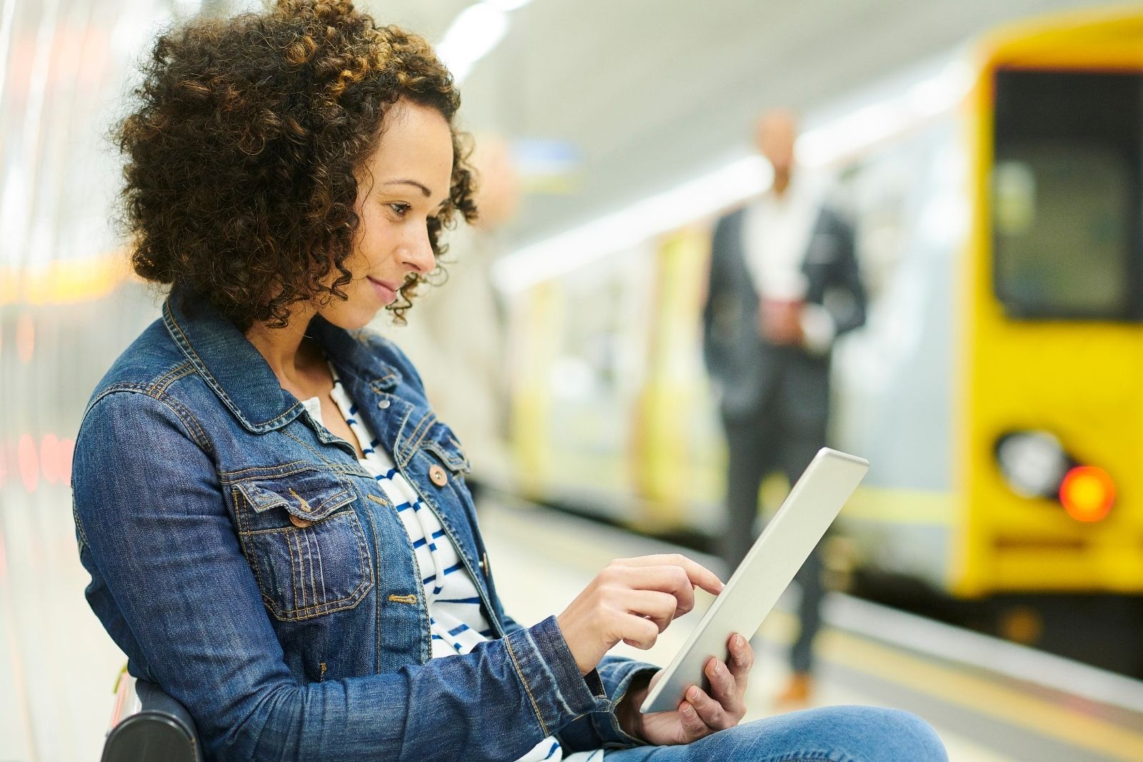 Young woman sits on metro platform reading book on her e-reader