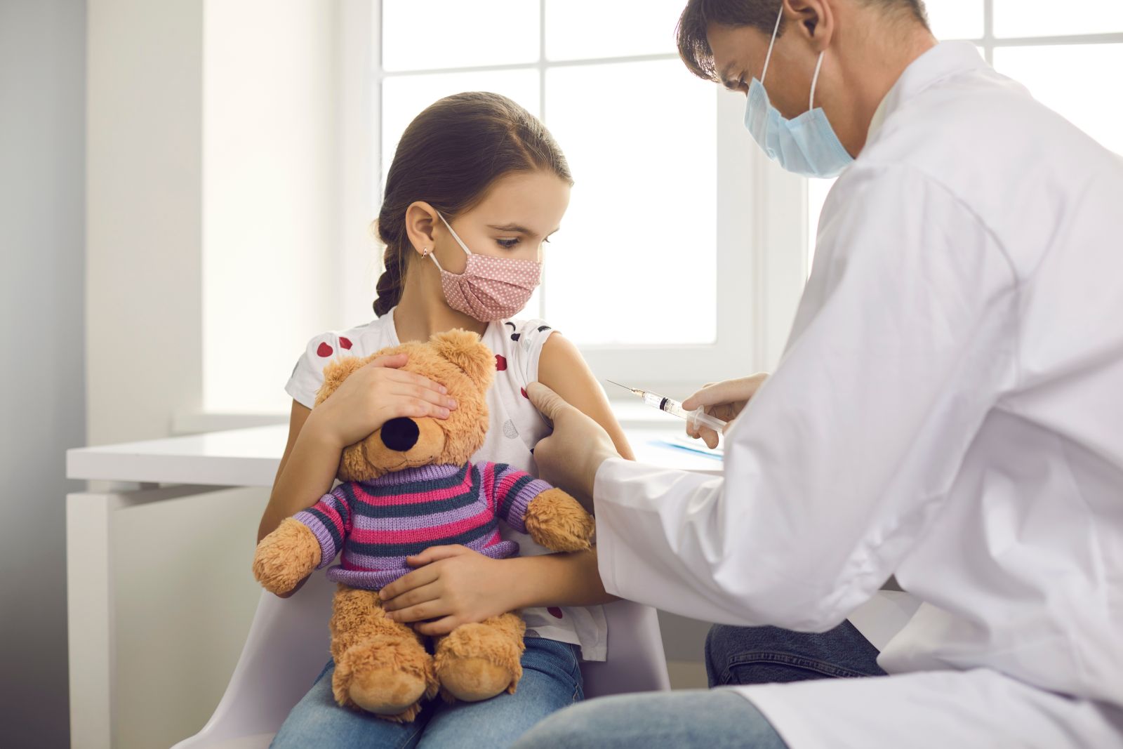 Young girl wearing facemask and holding teddy receives vaccination from doctor