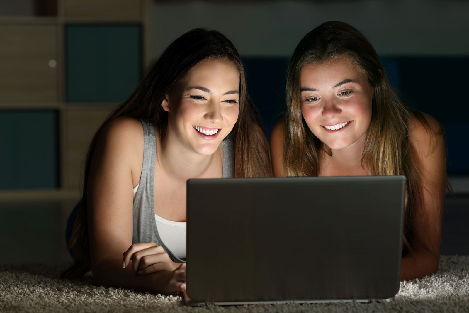 Two teens watching on line content in a laptop lying on the floor in a room at home with a dark light in the background