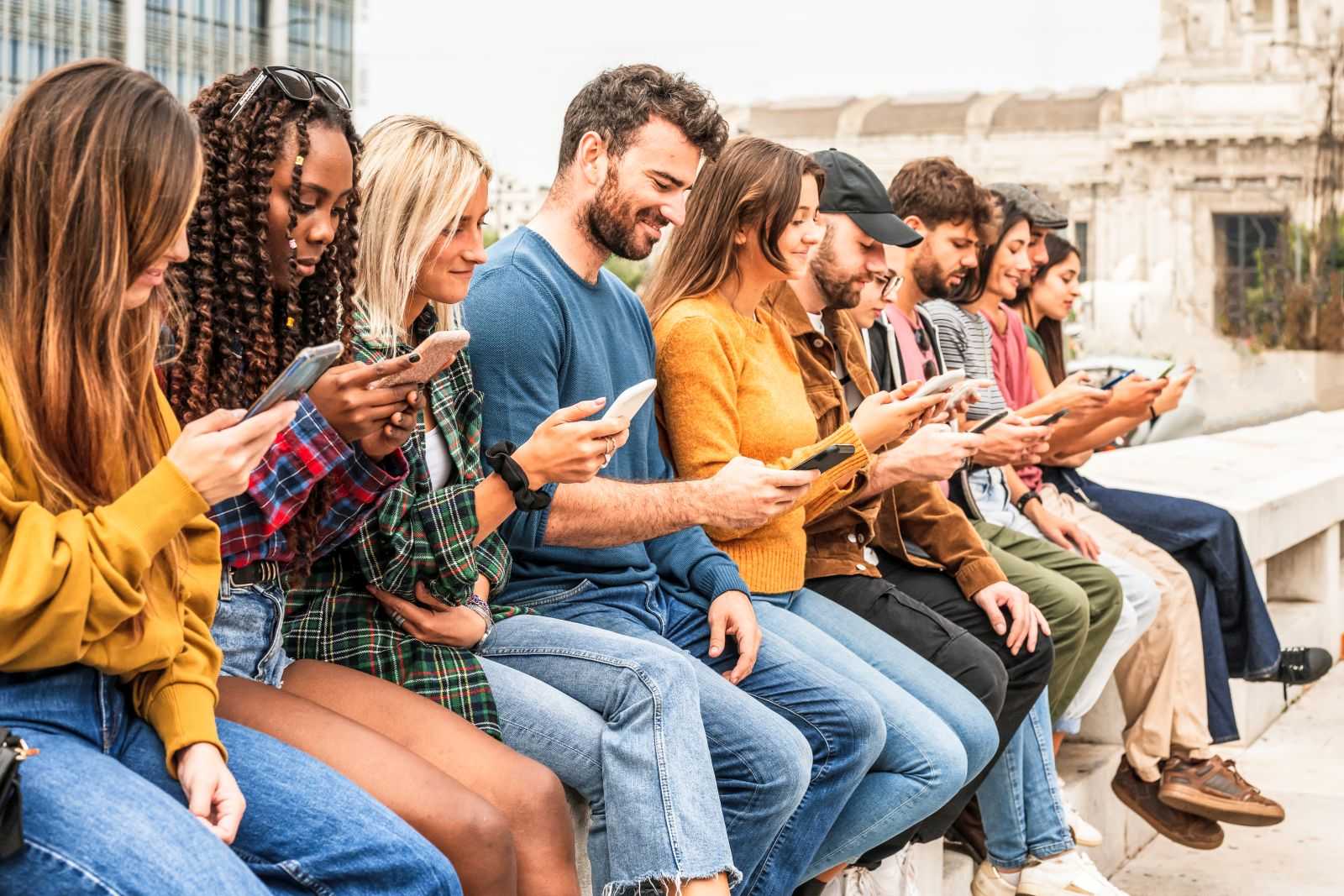 Group of young people sitting on a long bench outside reading news on their smartphones