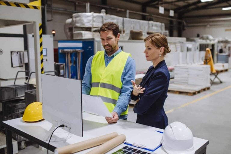 Facilities manager wearing protective gear talking to engineer in a manufacturing plant showing results on a computer