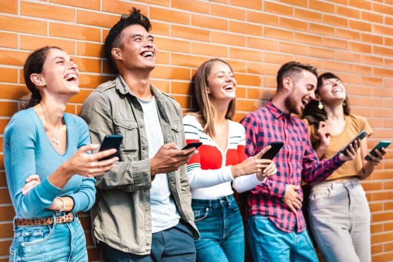 A group of young people lean against a brick wall smiling and laughing as they read something on their smartphones
