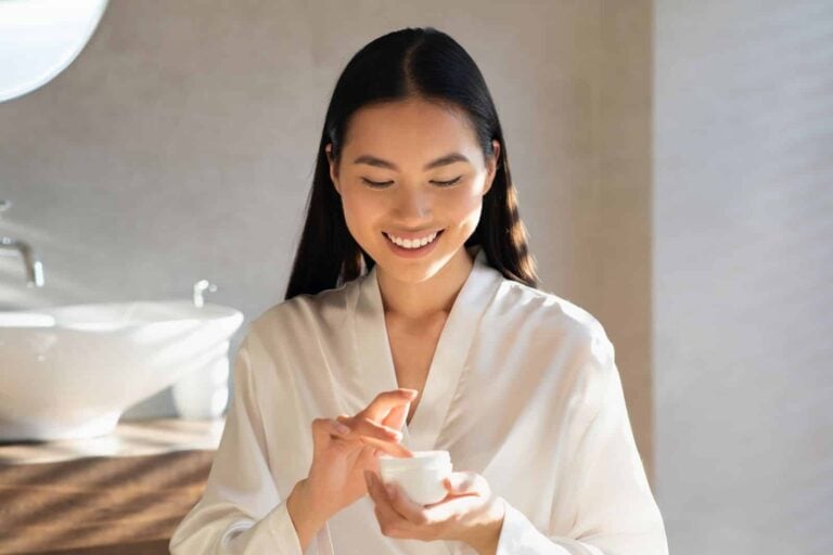 Smiling young asian lady in white bathrobe holding jar with beauty product, enjoying morning routine for beautiful face skin, standing in modern bathroom