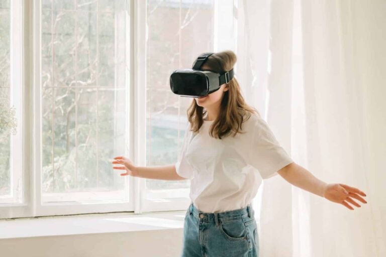 Young woman in white t-shirt and blue jeans plays game, dances with virtual reality headset helmet by the window in her room. Concept of entertainment, learning at home during quarantine