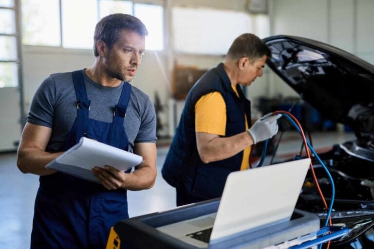 Two mechanics examining vehicle breakdown in auto repair shop. Focus is on mechanic using laptop while writing notes on clipboard.
