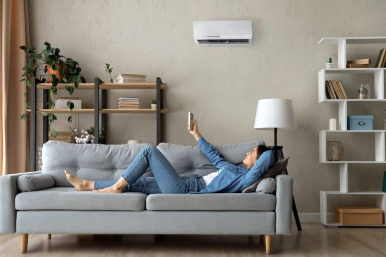 Relaxed young woman lying on sofa at home turning on air conditioner with remote control
