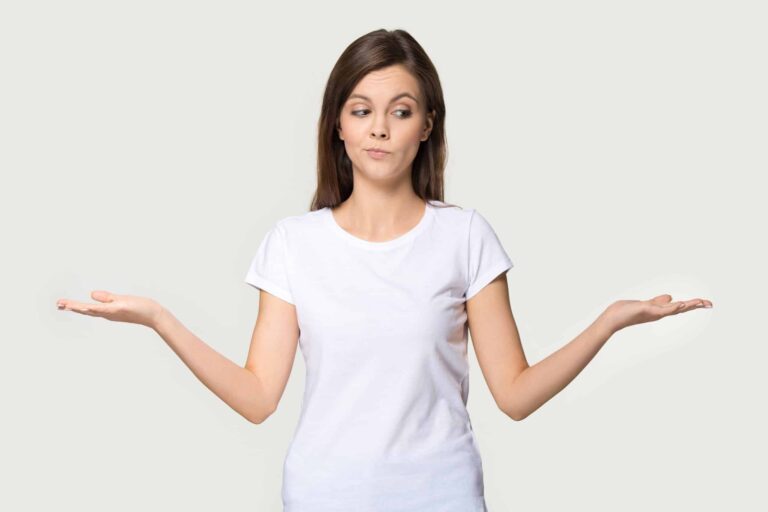 Young woman wearing white T-shirt stands looking confused and holding her arms out to the side with palms upwards