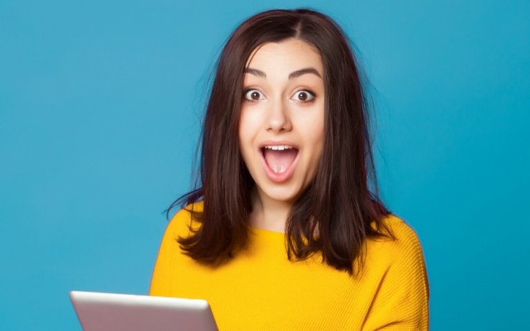 A surprised-looking young woman holds a tablet computer while standing against a blue background