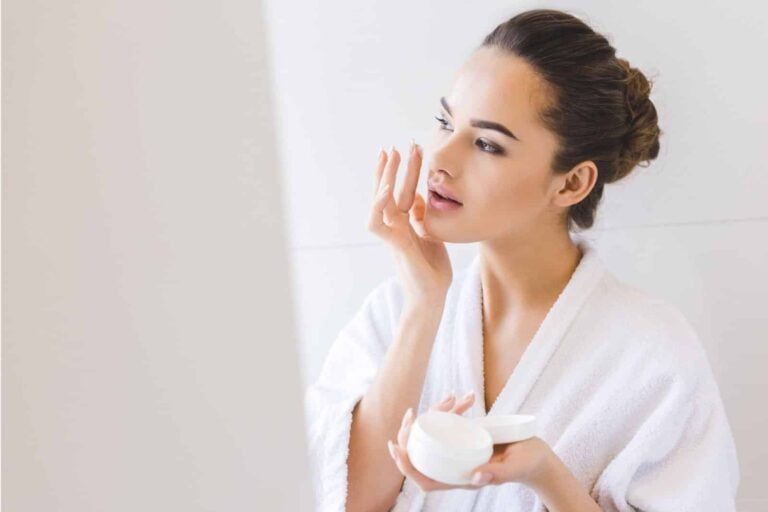 woman in white robe applying face cream while looking in a mirror
