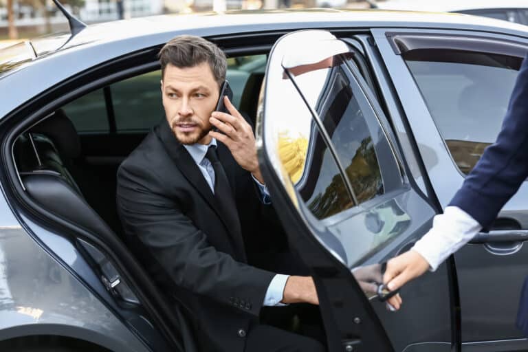 high net worth businessman in suit stepping out of car in the city using banking app on his phone