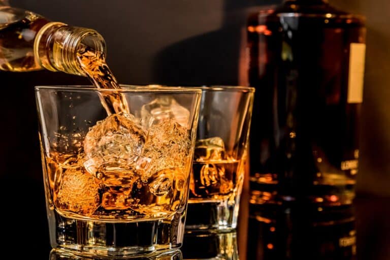 Pouring flavouired whiskey from a bottle into a glass containing ice. A second glass of whiskey and a bottom are visible in the background.