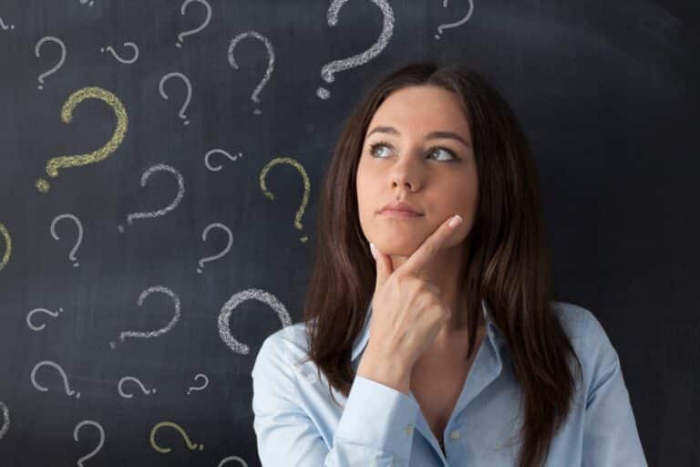 A business woman looking contemplative while standing in front of a blackboard with question marks on it