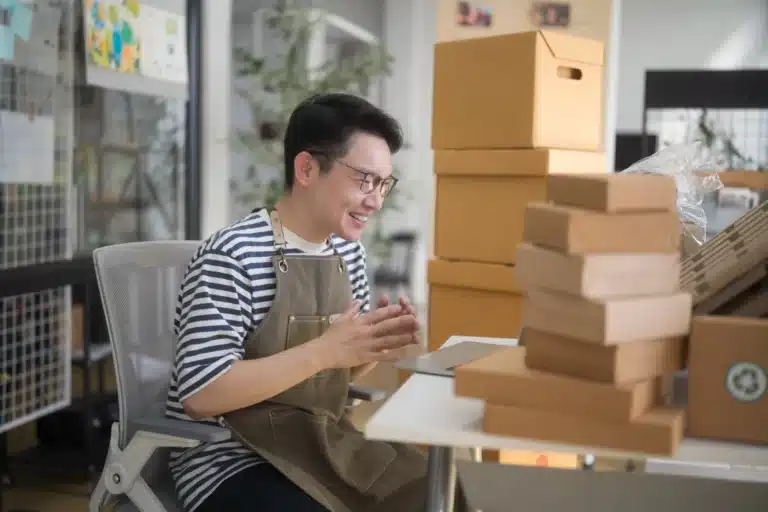 Smiling Vietnamese businessman taking part in interview about online advertising via a laptop while sitting at a desk surrounded by e-commerce parcels